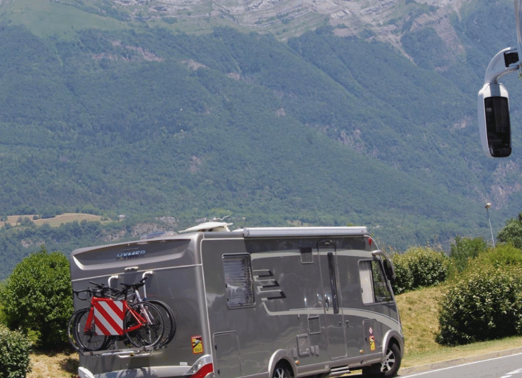 A grey motorhome driving along a road with two bicycles secured on the rear, set against a backdrop of green mountains.