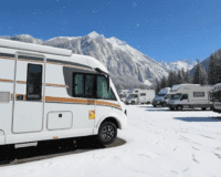 Close-up of a large white motorhome in a snowy parking area, with more campervans and snow-capped mountains visible in the background.