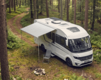 A high-angle shot of a modern, white recreational vehicle (RV) parked on a dirt path in the middle of a dense, lush green forest. The RV has its side awning extended, sheltering a single, empty gray folding chair next to a small rock campfire ring on the mossy ground. Tall pine and deciduous trees with thick foliage surround the clearing, creating a serene, secluded camping atmosphere.