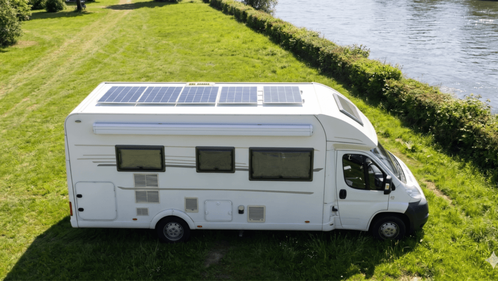A high-angle, side-profile view of a white motorhome parked on a vibrant green, grassy bank next to a wide, sunlit river. The camper van features large solar panels covering its roof, tinted windows, a side awning, and a white cab with a black bumper. The background shows a long, manicured green hedge separating the grassy area from the water, with lush green trees visible in the far upper-left corner under bright, natural daylight.