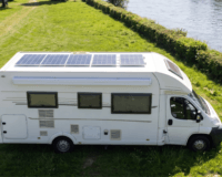 A high-angle, side-profile view of a white motorhome parked on a vibrant green, grassy bank next to a wide, sunlit river. The camper van features large solar panels covering its roof, tinted windows, a side awning, and a white cab with a black bumper. The background shows a long, manicured green hedge separating the grassy area from the water, with lush green trees visible in the far upper-left corner under bright, natural daylight.