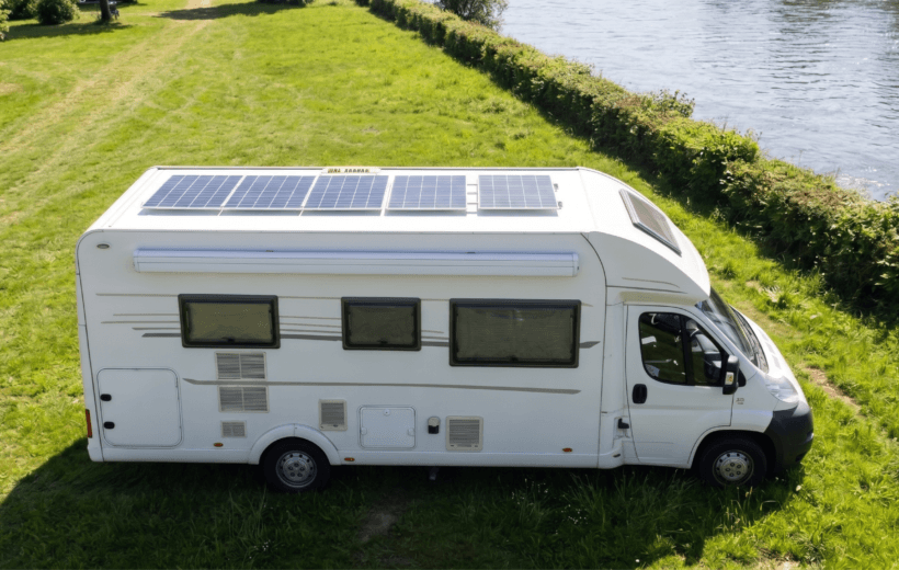 A high-angle, side-profile view of a white motorhome parked on a vibrant green, grassy bank next to a wide, sunlit river. The camper van features large solar panels covering its roof, tinted windows, a side awning, and a white cab with a black bumper. The background shows a long, manicured green hedge separating the grassy area from the water, with lush green trees visible in the far upper-left corner under bright, natural daylight.