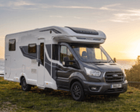 white and grey Ford Bailey Adamo motorhome parked on a grassy clifftop overlooking the sea during a bright sunset. The motorhome is positioned at a three-quarter angle facing right, showing its distinct dark grey front cab with a Ford grille and a British "BAILEY" license plate.