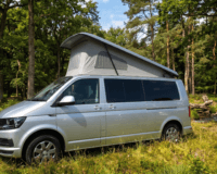 A silver Volkswagen danbury camper van is parked in a lush forest clearing.